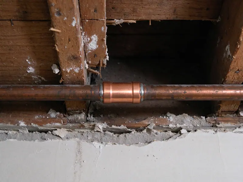 Close view of a new bare copper repair coupling soldered into an old copper water line inside an opened ceiling between wooden joists, the joint clean with a thin line of solder