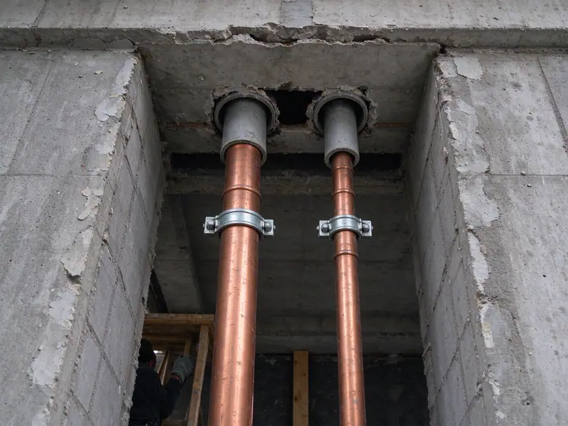Looking up into an opened chase in an apartment building showing new bare copper water risers with riser clamps passing through a sleeve in the concrete floor slab above