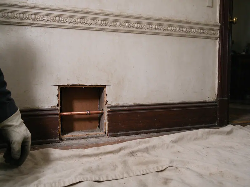 A neat square access opening cut in an old plaster parlor floor baseboard area of a Brooklyn brownstone, ornate plaster molding above it, showing new copper pipe inside